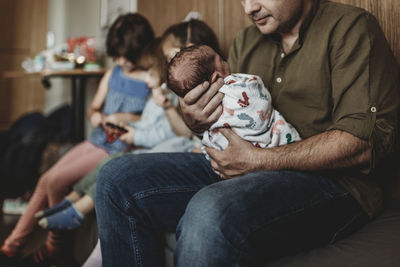 Side view of father holding newborn with siblings in background