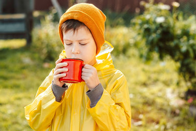 A little boy in a yellow raincoat is drinking from a red mug on the street. camping concept. 