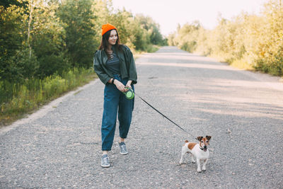 Cute modern teen girl in a green jacket and orange hat walks with her dog in nature. 