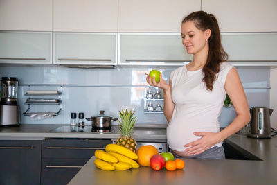 Pregnant woman holding fruit standing in kitchen at home