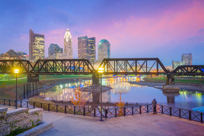 Illuminated bridge over river by buildings against sky at sunset