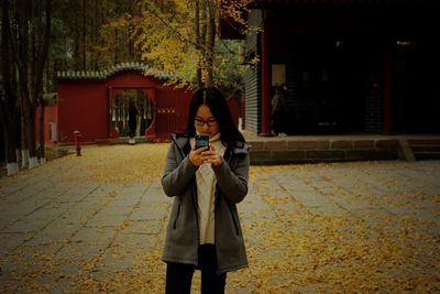 Young woman photographing with mobile phone while standing at park during autumn