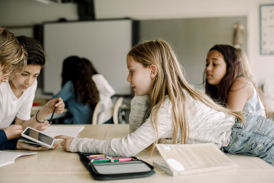 Female student pointing at digital tablet while leaning over table in classroom