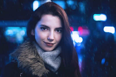 Portrait of young woman smiling while standing in illuminated city at night during snowfall