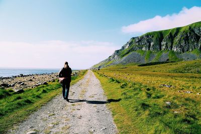 Full length rear view of traveler walking on dirt road at sea shore by mountain against sky