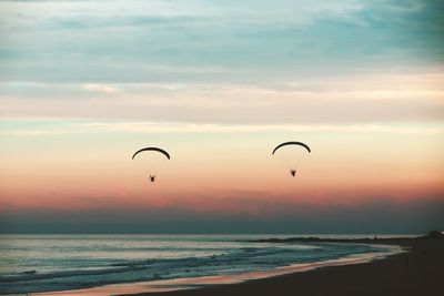 Silhouette parachutes flying over sea against orange sky