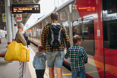 Back shot of caucasian man holding children hands walking along platform going to take train