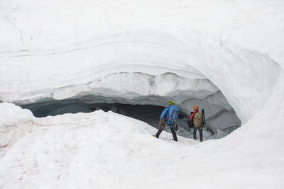 Two explorers enter the mouth of a glacial cave with gear in hand.