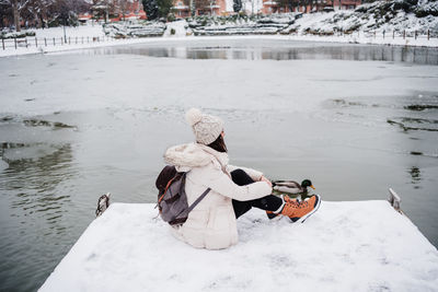 Back view of backpacker woman sitting on pier in front of frozen lake in city. 