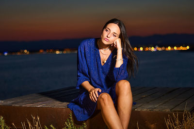 Beautiful young woman sitting against sky during sunset