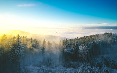 Wind turbine behind a forest covered in fog during a colorful sunset.