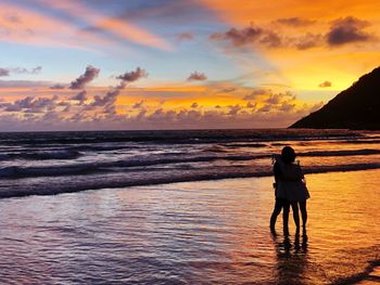 Silhouette couple standing at beach against dramatic sky during sunset