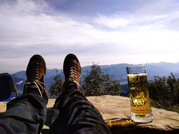 Low section of man resting by beer glass on rock against sky