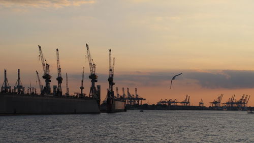 Cranes at commercial dock against sky during sunset