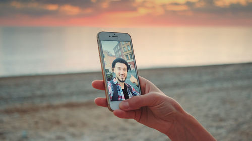 Cropped hand of woman holding mobile phone while video conferencing with man at beach