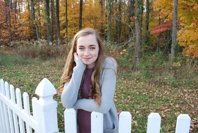 Portrait of smiling young woman standing by fence against trees in forest during autumn