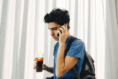 Young man answering smart phone while holding cola in restaurant