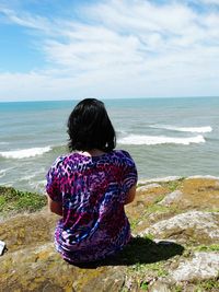 Rear view of woman sitting on rock formation by sea against sky