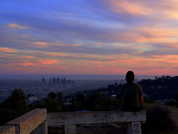 Rear view of man looking at cityscape against sky during sunset
