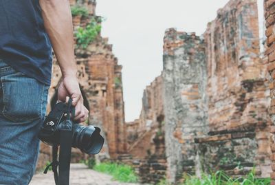 Midsection of man holding camera while standing against stone wall