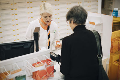 Rear view of senior customer standing with female owner at checkout in pharmacy store