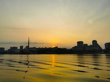 Silhouette buildings by river against sky during sunset