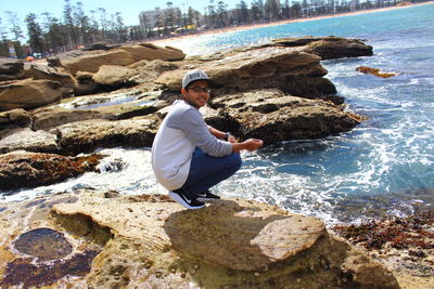 Full length portrait of man crouching on rock at beach