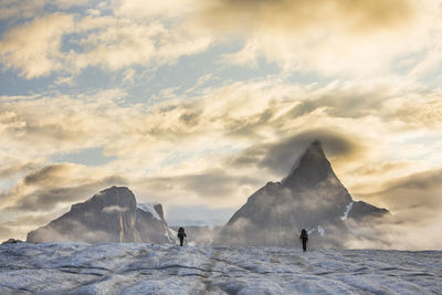 Silhouette view of two backpackers hiking on glacier to mountains.