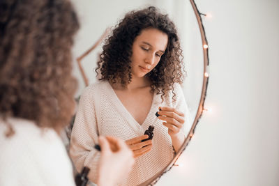 Young woman doing self-care routine at mirror, using aroma oil or serum for skin