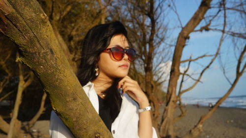 Young woman wearing sunglasses by tree trunk at beach