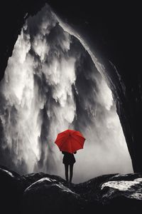 Rear view of person holding umbrella while standing on rock against waterfall