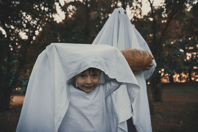 Portrait of cute boy with person wearing ghost costume in park at sunset during halloween