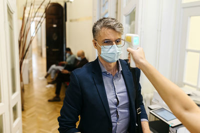 Hand of female with infrared thermometer checking mature businessman's temperature in corridor