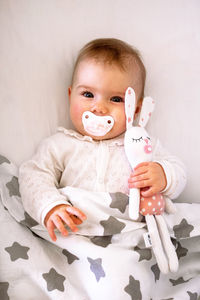 Candid portrait of smiling baby girl lying on bed with pacifier and toy of hare in her hand.