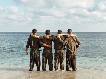 Rear view of army soldiers standing with arms around in sea against cloudy sky