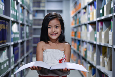 Portrait of smiling girl holding book while standing in library