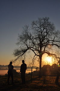 Silhouette people standing by bare trees against sky during sunset