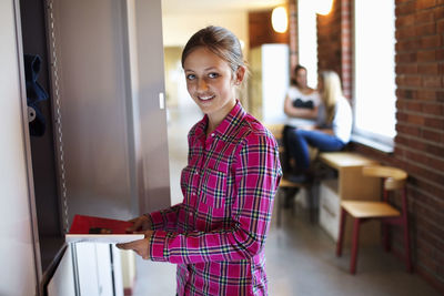 Portrait of schoolgirl holding book while standing by locker