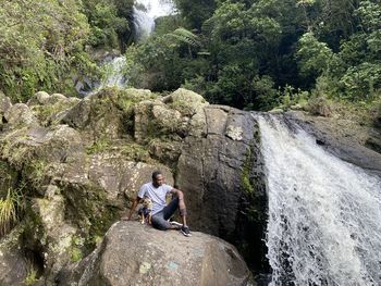 Front view of man and fox terrier pug dog sitting on rock infront of waterfall