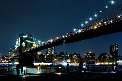 Illuminated brooklyn bridge over river against buildings and sky in city at night