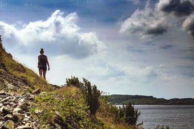 Rear view of young woman standing on field by sea against sky