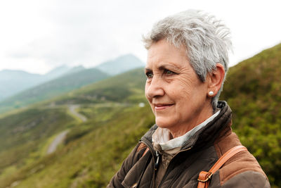 Positive elderly female traveler face and looking away on blurred background of nature