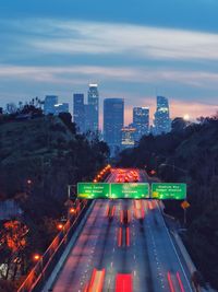 Illuminated city buildings against sky at dusk and highway light trails