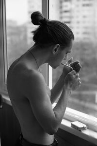 Shirtless young man smoking cigarette by window at home