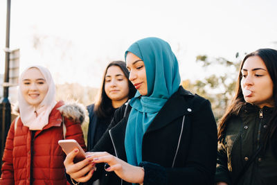 Young muslim woman using smart phone while standing with female friends against sky