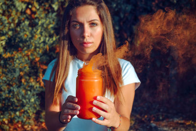 Portrait of woman holding jar with orange smoke while standing against plants