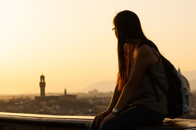 Woman sitting on building terrace in city against sky during sunset
