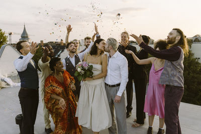 Happy wedding guests throwing confetti on newlywed couple kissing on rooftop during wedding celebration