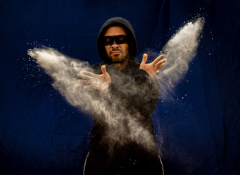 Man wearing hood with face paint throwing powder against backdrop
