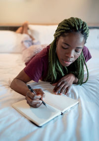 Concentrated young african american woman in pajama resting on bed and writing in diary in modern bedroom at home